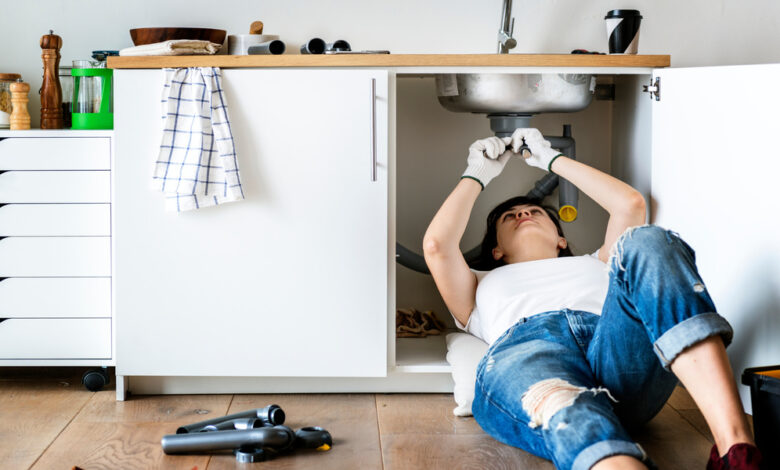 A woman wearing ripped jeans and a white t-shirt lays under her kitchen sink to repair it.