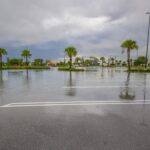 An empty parking lot with palm trees is flooded with water pooled on the asphalt under a sky with lots of clouds.