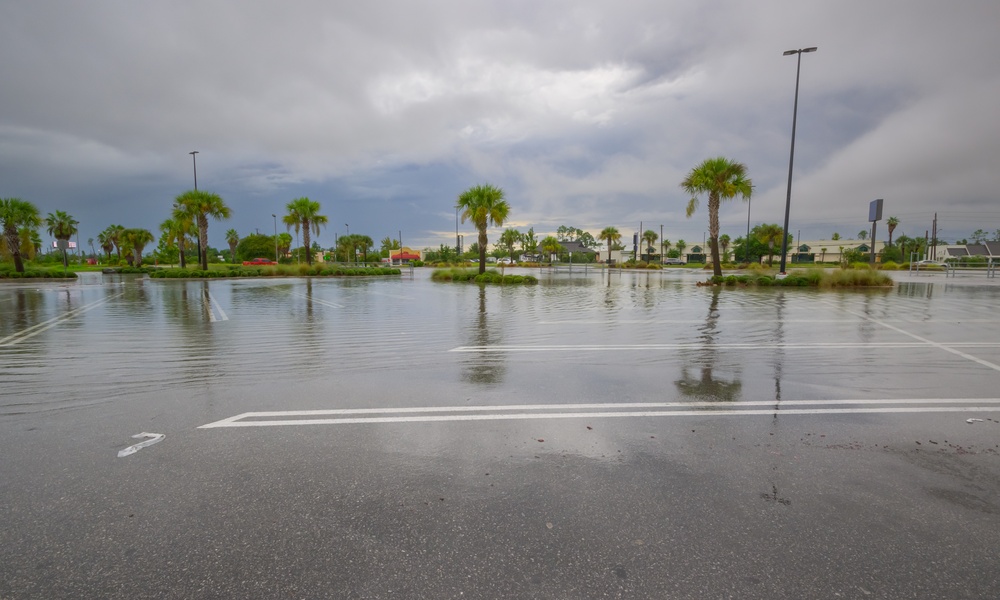 An empty parking lot with palm trees is flooded with water pooled on the asphalt under a sky with lots of clouds.