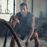 A young man in the gym using ropes to work out his arms. He's moving them up and down and there's steam around him.