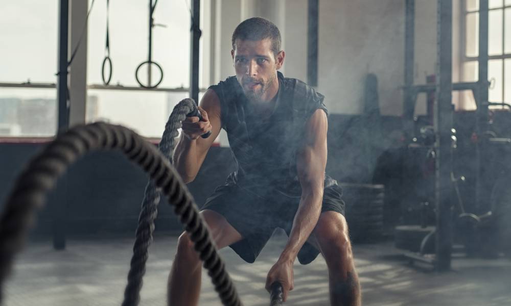 A young man in the gym using ropes to work out his arms. He's moving them up and down and there's steam around him.