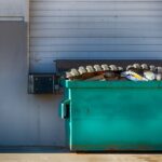 A large green dumpster sitting outside of a business docking station. The dumpster is full of garbage and waste.