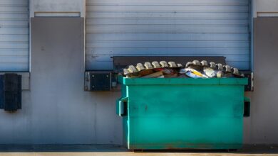 A large green dumpster sitting outside of a business docking station. The dumpster is full of garbage and waste.