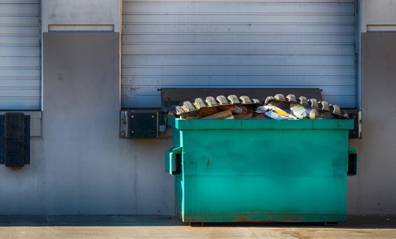 A large green dumpster sitting outside of a business docking station. The dumpster is full of garbage and waste.