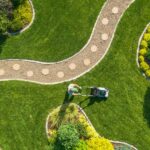 A man with a green hat pushing a lawn mower across a large green lawn with a curved gravel path through it.