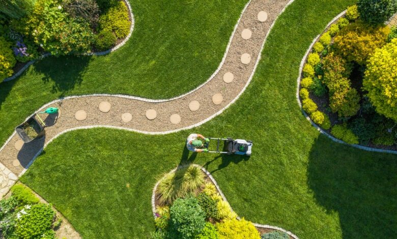 A man with a green hat pushing a lawn mower across a large green lawn with a curved gravel path through it.