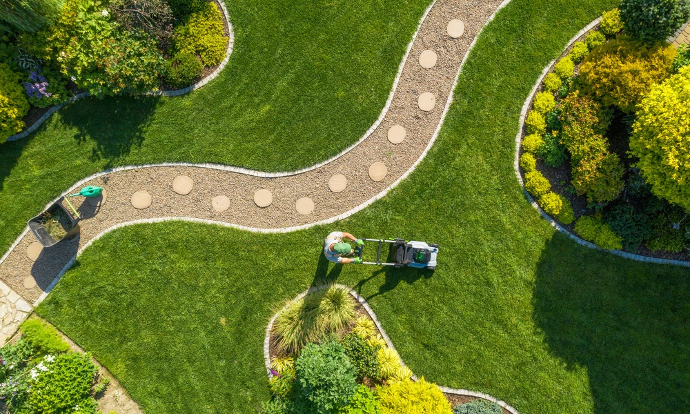 A man with a green hat pushing a lawn mower across a large green lawn with a curved gravel path through it.