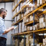 A man in a blue shirt stands and looks at a tablet in his hands in front of a large warehouse pallet rack unit.