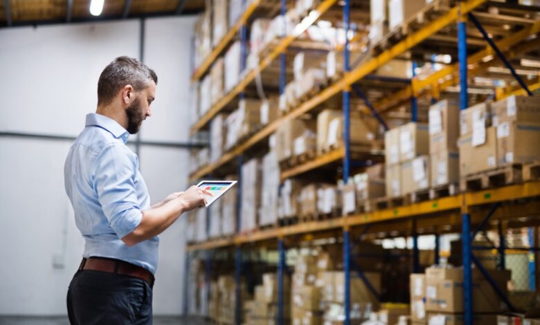 A man in a blue shirt stands and looks at a tablet in his hands in front of a large warehouse pallet rack unit.