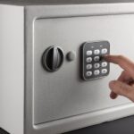 An individual is using their finger to type in a numerical code on the keypad of a small gray safe on a black background.