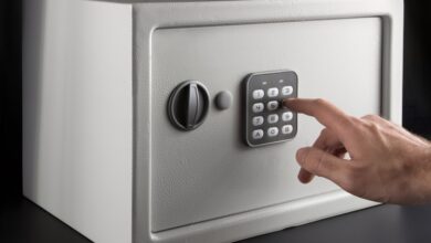 An individual is using their finger to type in a numerical code on the keypad of a small gray safe on a black background.
