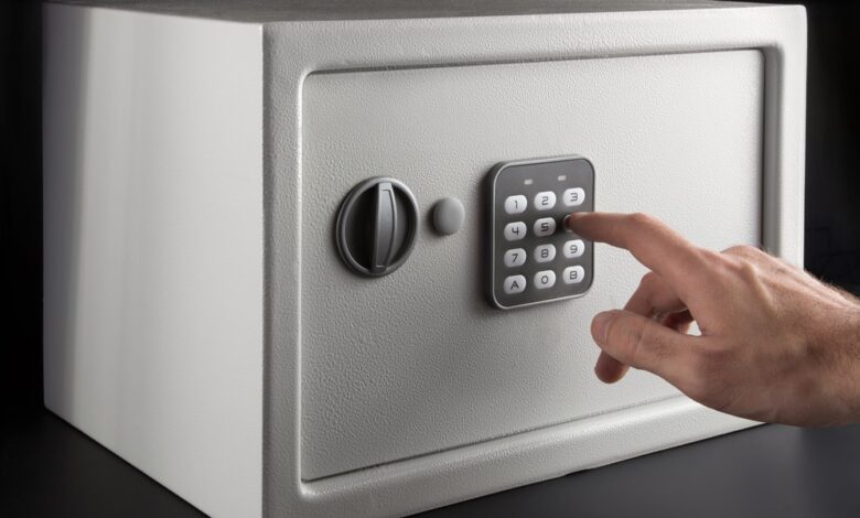 An individual is using their finger to type in a numerical code on the keypad of a small gray safe on a black background.