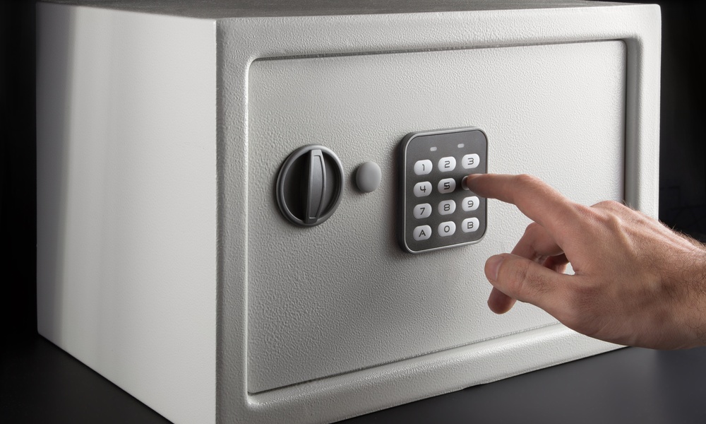 An individual is using their finger to type in a numerical code on the keypad of a small gray safe on a black background.