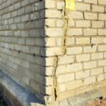 At the outer corner of a brick house, one wall has a long crack going up the side. The house sits on a brick foundation.