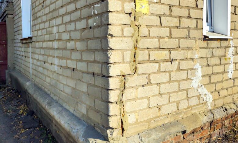 At the outer corner of a brick house, one wall has a long crack going up the side. The house sits on a brick foundation.