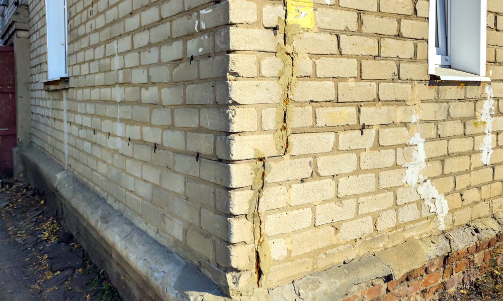 At the outer corner of a brick house, one wall has a long crack going up the side. The house sits on a brick foundation.