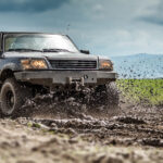 A car is driving off-road through a large patch of mud that flies all over. Behind the car is a gray sky and hills.