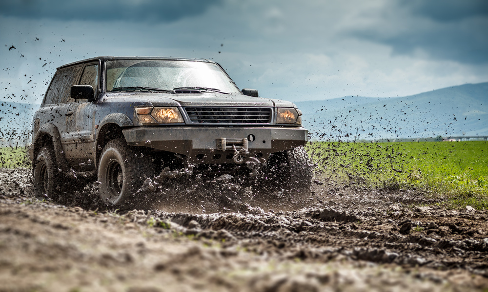 A car is driving off-road through a large patch of mud that flies all over. Behind the car is a gray sky and hills.