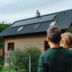 A man wearing a green sweater holds a small child as they look at a house that has solar panels on the roof.