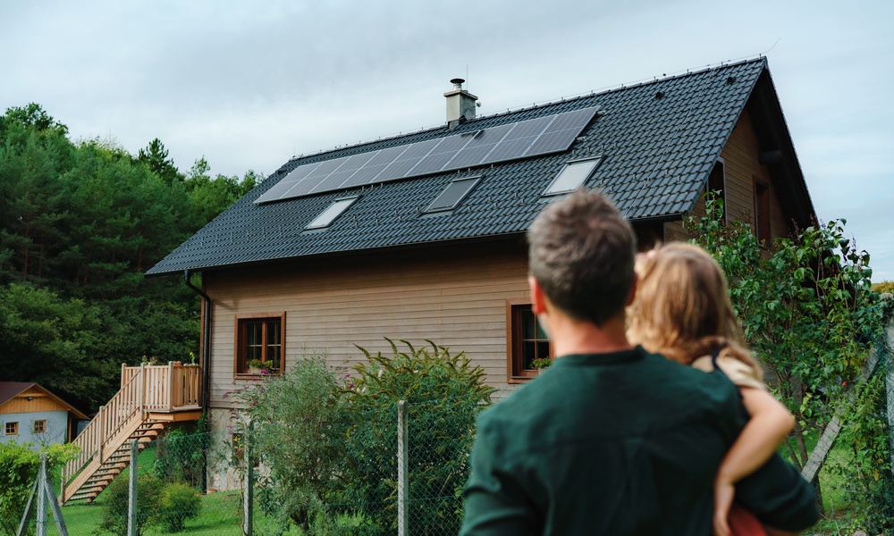 A man wearing a green sweater holds a small child as they look at a house that has solar panels on the roof.