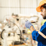 A man wearing an orange hard hat and a blue protective suit with reflective strips is examining a piece of metal.