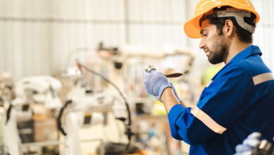 A man wearing an orange hard hat and a blue protective suit with reflective strips is examining a piece of metal.