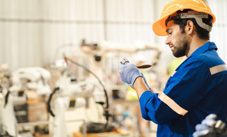A man wearing an orange hard hat and a blue protective suit with reflective strips is examining a piece of metal.