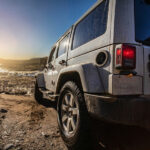 Rear view of a Jeep as it goes off-roading on a dirt road. The sun hangs in the sky in the background, shining bright.