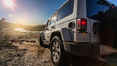 Rear view of a Jeep as it goes off-roading on a dirt road. The sun hangs in the sky in the background, shining bright.