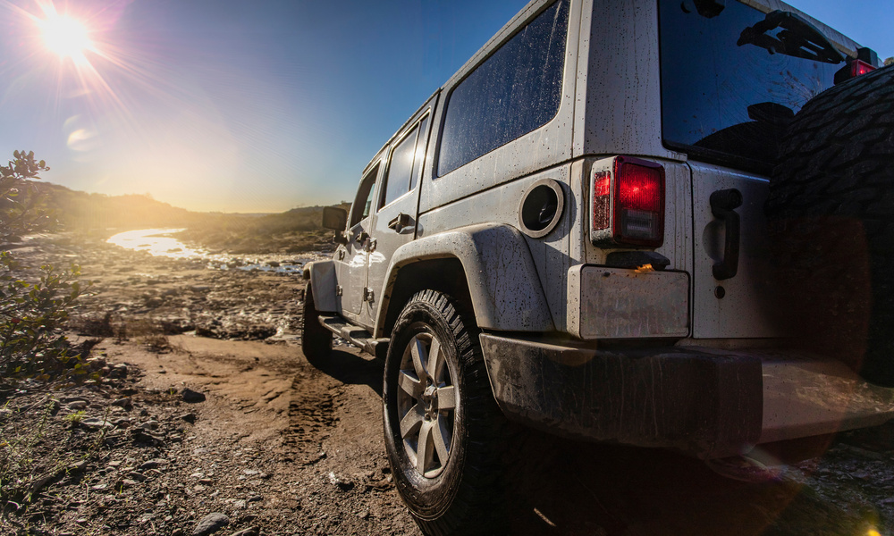 Rear view of a Jeep as it goes off-roading on a dirt road. The sun hangs in the sky in the background, shining bright.