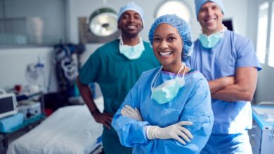 A smiling female healthcare professional crosses her arms across her chest and stands in front of two male surgeons.