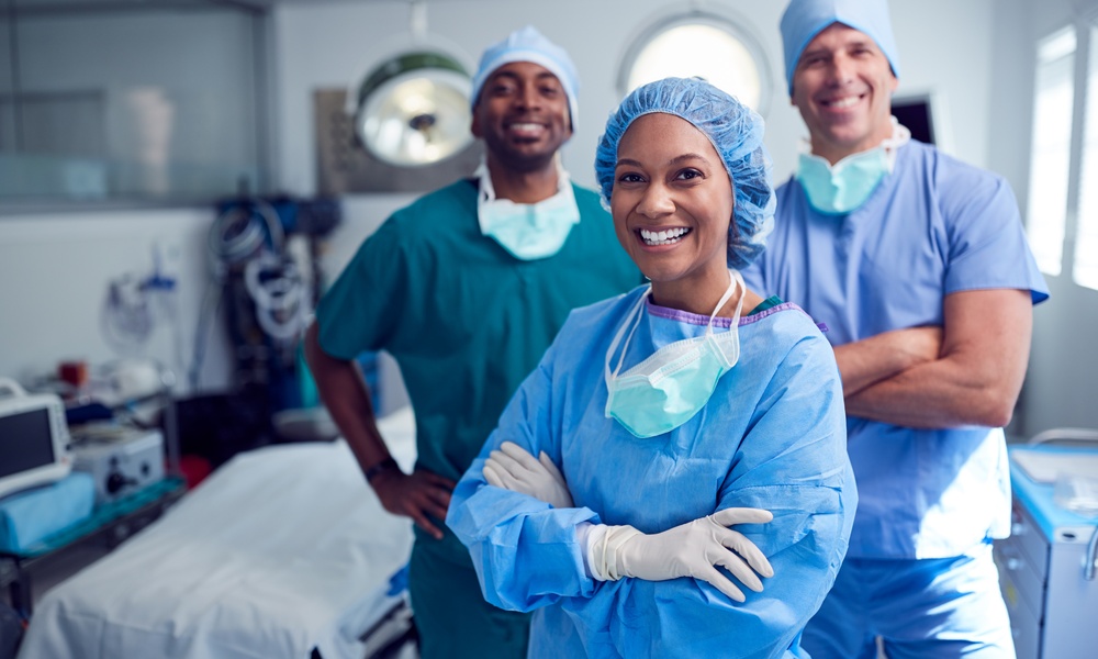 A smiling female healthcare professional crosses her arms across her chest and stands in front of two male surgeons.