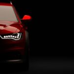 A stylish red car against a black background, part of the car hidden in shadows. The headlights are on.