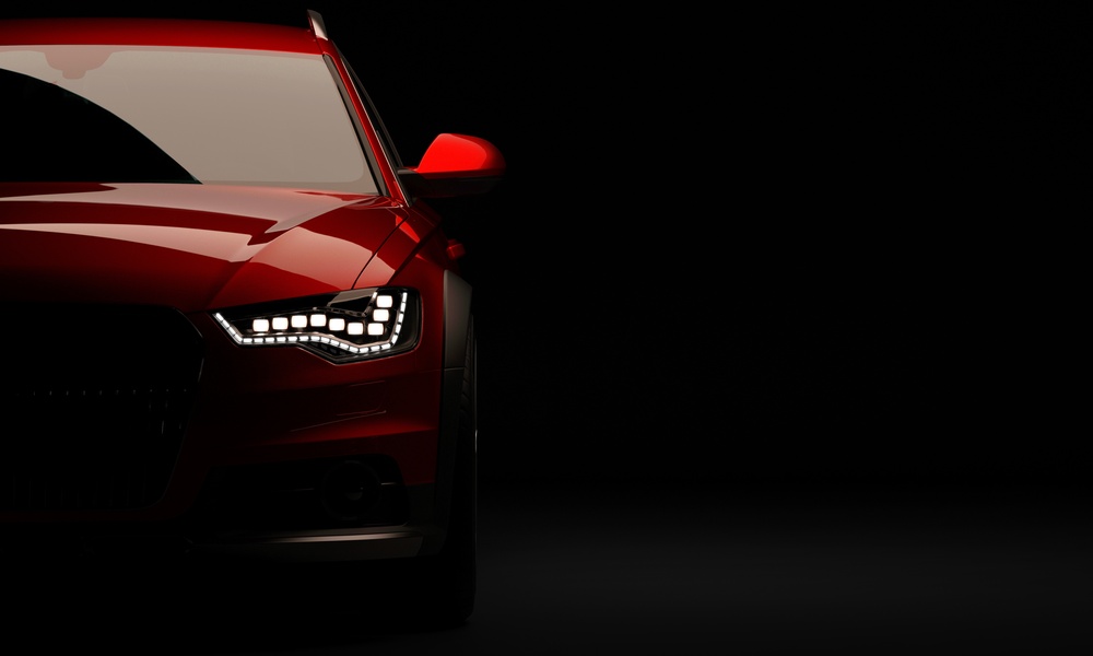 A stylish red car against a black background, part of the car hidden in shadows. The headlights are on.
