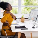 A woman sitting at a desk in an ergonomic chair in a home office. She's rubbing her shoulder and lower back.