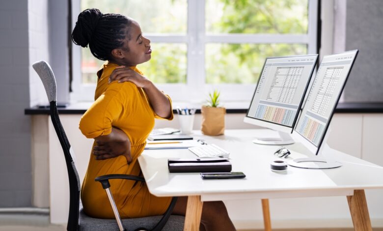 A woman sitting at a desk in an ergonomic chair in a home office. She's rubbing her shoulder and lower back.