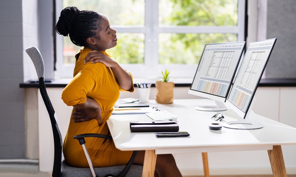 A woman sitting at a desk in an ergonomic chair in a home office. She's rubbing her shoulder and lower back.