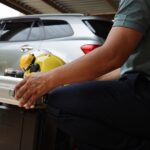 A man in jeans squats down and holds a metal case. There is a yellow hard hat and ear protectors on top.