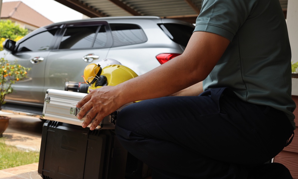 A man in jeans squats down and holds a metal case. There is a yellow hard hat and ear protectors on top.