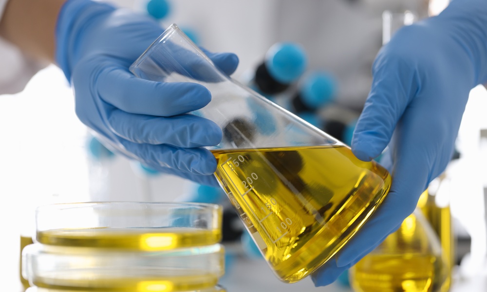 A pair of hands wearing light blue latex gloves is carefully holding an Erlenmeyer flask filled with bright yellow liquid.
