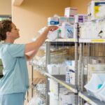 A nurse in light blue scrubs reaches into a box on the top shelf of a shelving unit holding various medical supplies.