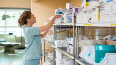 A nurse in light blue scrubs reaches into a box on the top shelf of a shelving unit holding various medical supplies.