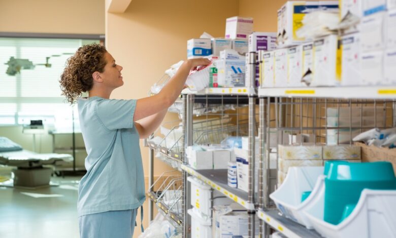 A nurse in light blue scrubs reaches into a box on the top shelf of a shelving unit holding various medical supplies.