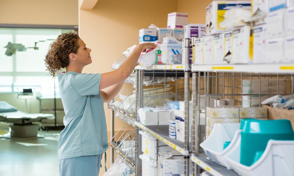A nurse in light blue scrubs reaches into a box on the top shelf of a shelving unit holding various medical supplies.
