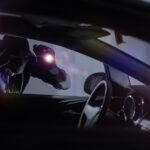A car thief wearing all black and a mask holds a flashlight to the window of a car, looking inside.