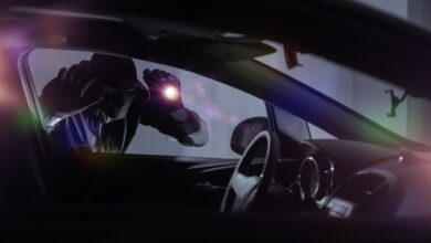 A car thief wearing all black and a mask holds a flashlight to the window of a car, looking inside.