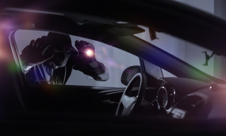 A car thief wearing all black and a mask holds a flashlight to the window of a car, looking inside.