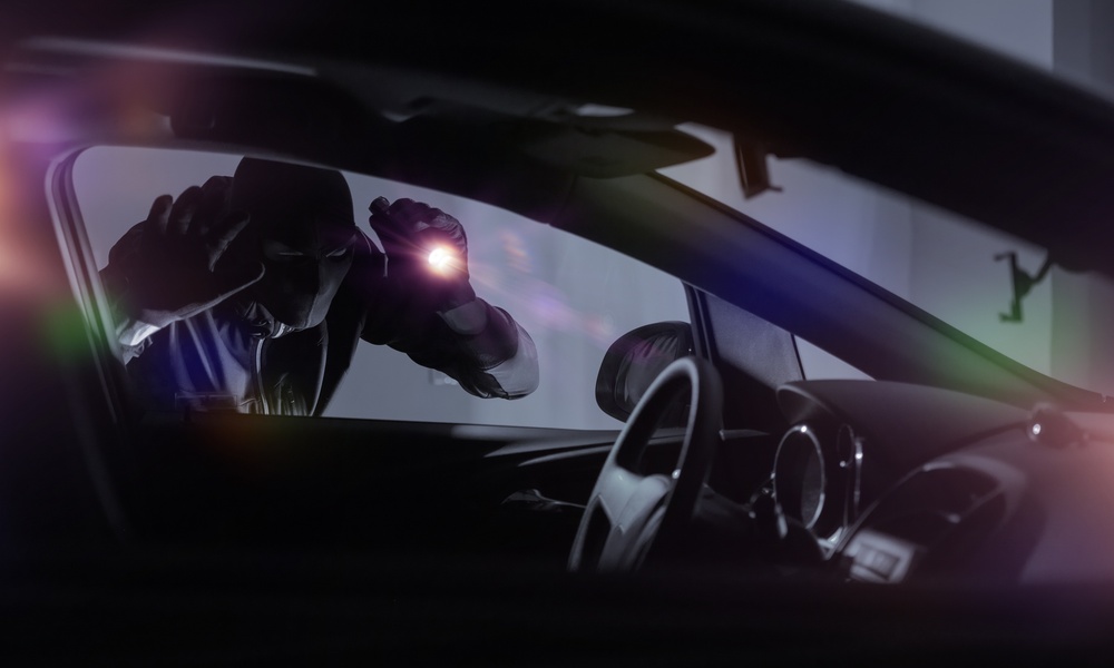 A car thief wearing all black and a mask holds a flashlight to the window of a car, looking inside.