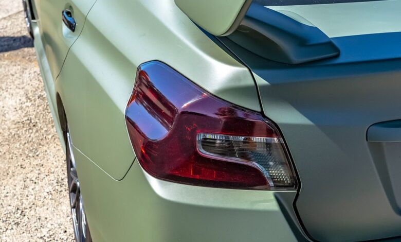 A close-up of the left taillight and spoiler of a vehicle. The car is matte green and parked on gravel.