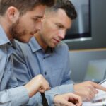 Two male technicians look at an equipment manual together. One man points at the handbook while the other looks on.